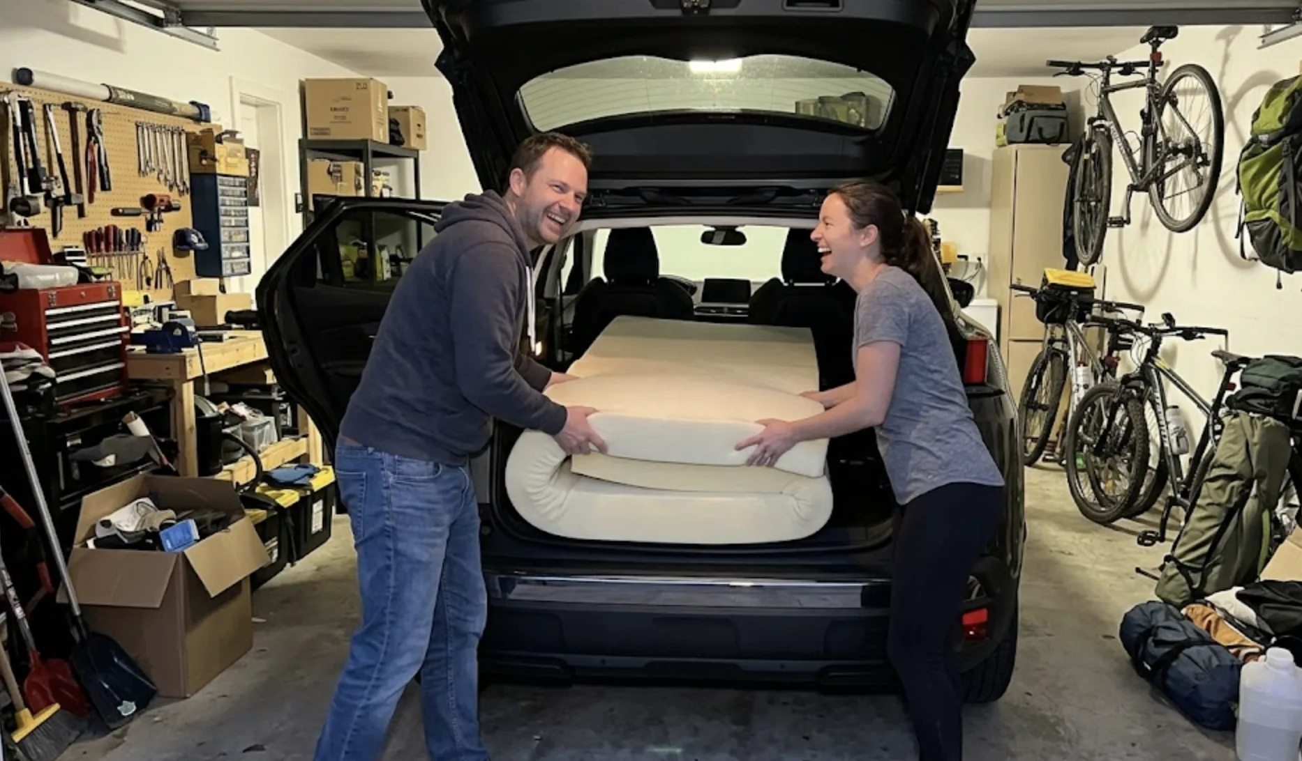 Deux personnes dans la trentaine en train d'installer un matelas en mousse plié dans le coffre d'un Peugeot 3008, lumière chaleureuse de fin d'après-midi dans un garage