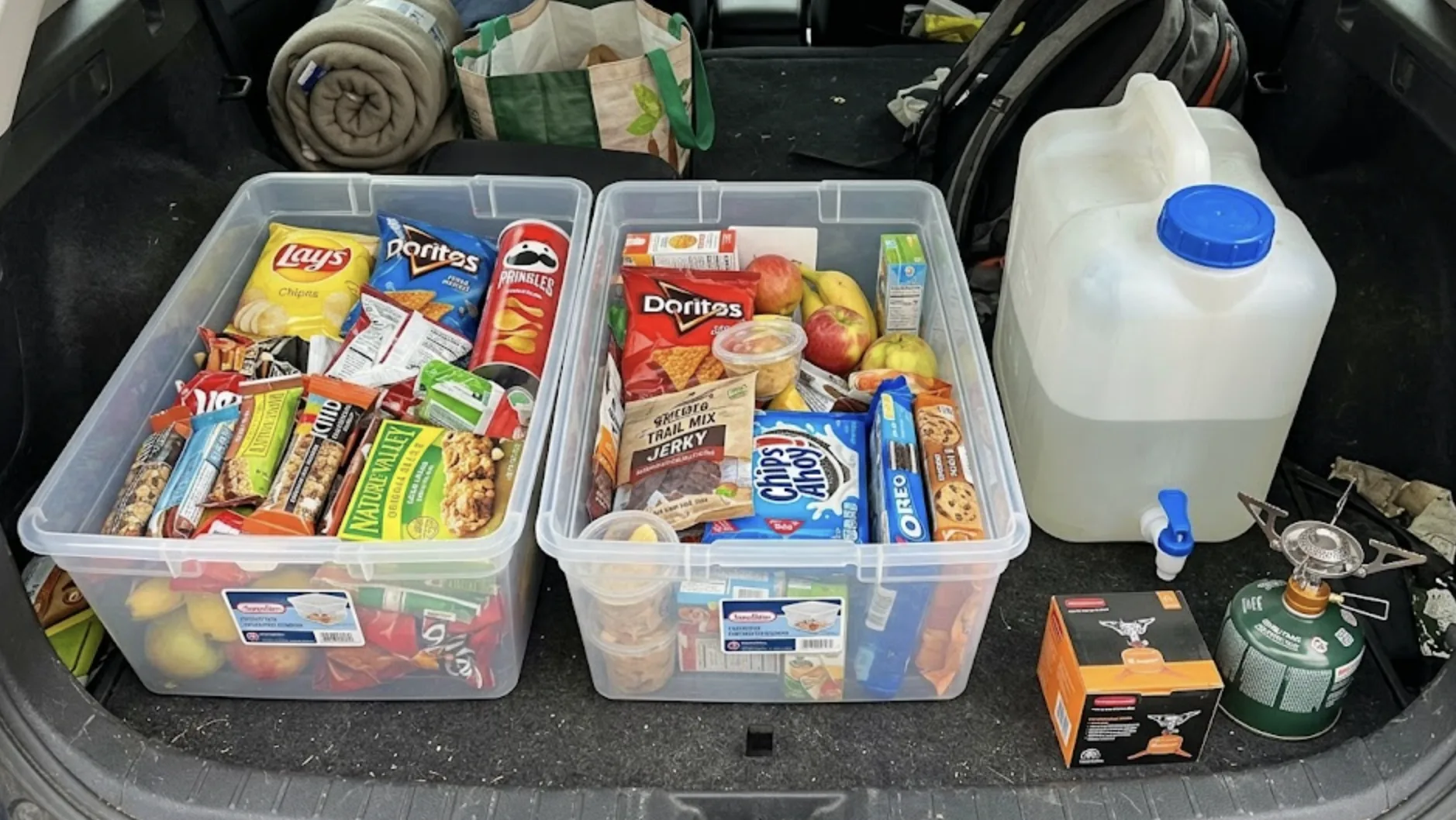 Gros plan dans un coffre de voiture : deux caisses en plastique transparent remplies de nourriture de road trip, un bidon d'eau de 10 litres avec robinet, et un petit réchaud à gaz portable — organisation soignée