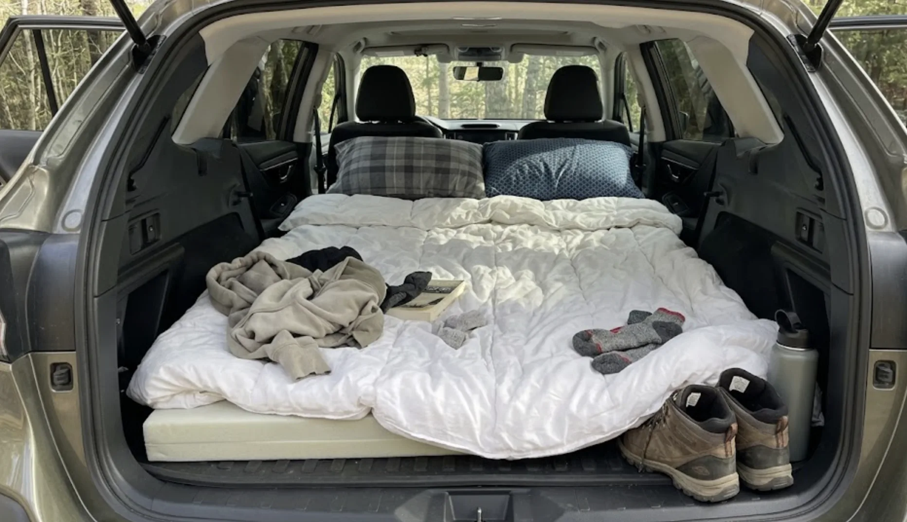 Setup de couchage douillet dans le coffre d'un SUV : matelas en mousse recouvert d'une épaisse couette blanche et deux oreillers à mémoire de forme — vue depuis le hayon ouvert, atmosphère chaleureuse