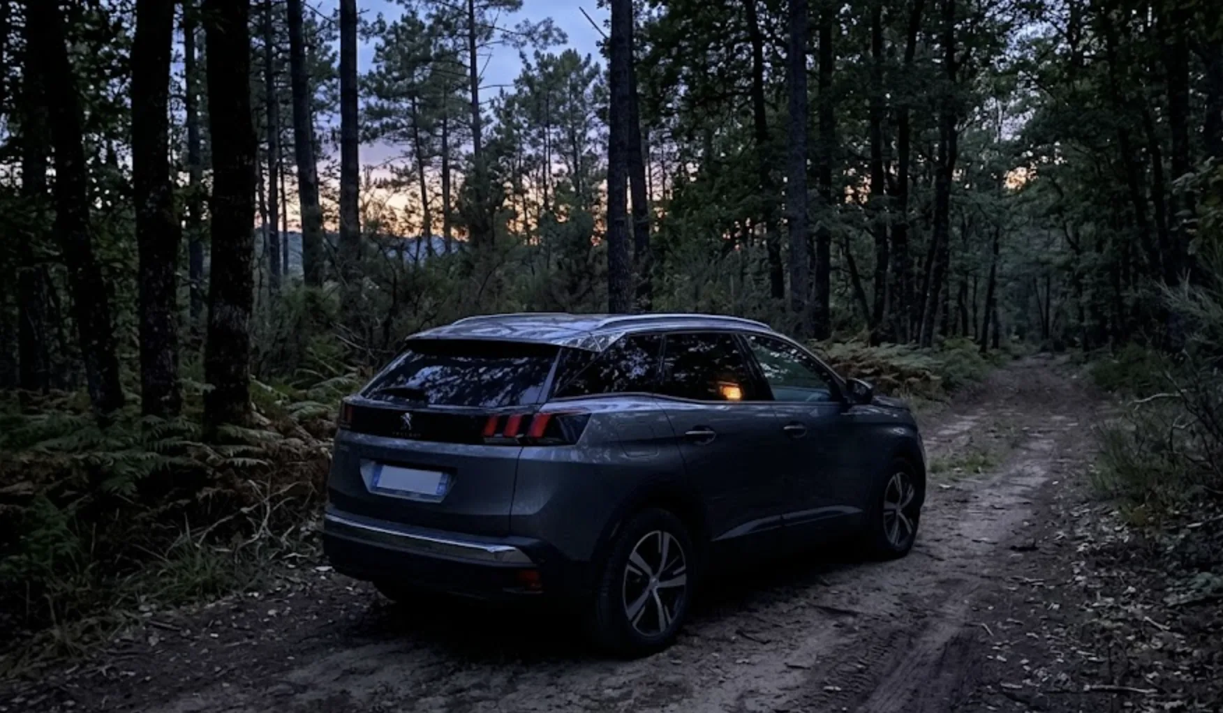 Peugeot 3008 garé sur un chemin de terre entouré d'arbres en Ardèche au crépuscule — la voiture semble banale de l'extérieur mais une faible lueur orangée chaleureuse est visible à travers les vitres teintées arrière