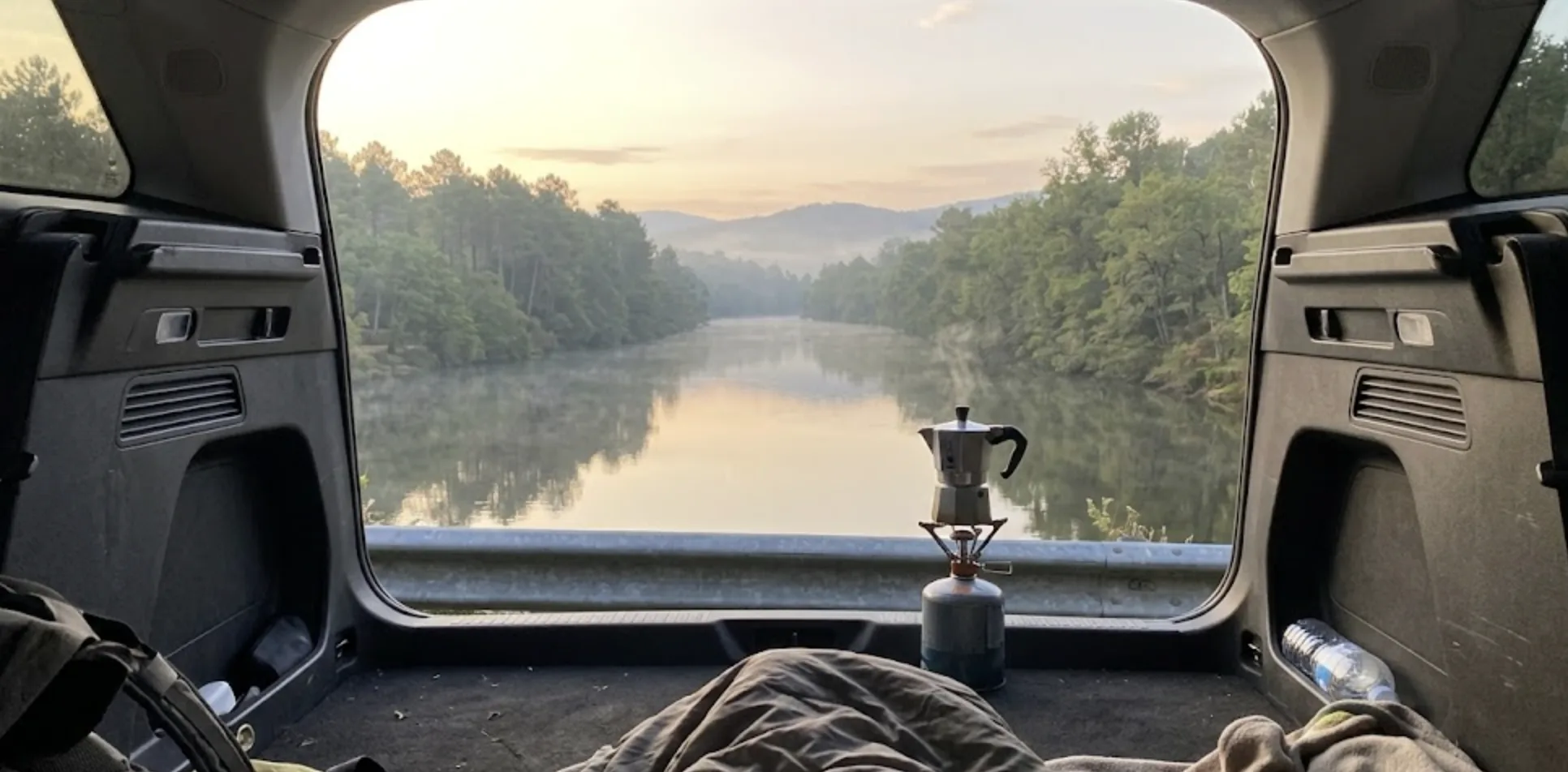Vue subjective au réveil depuis l'intérieur d'un coffre de voiture : hayon grand ouvert sur une rivière et des falaises rocheuses dans la brume matinale, au premier plan une cafetière italienne sur un réchaud de camping posé sur le pare-chocs