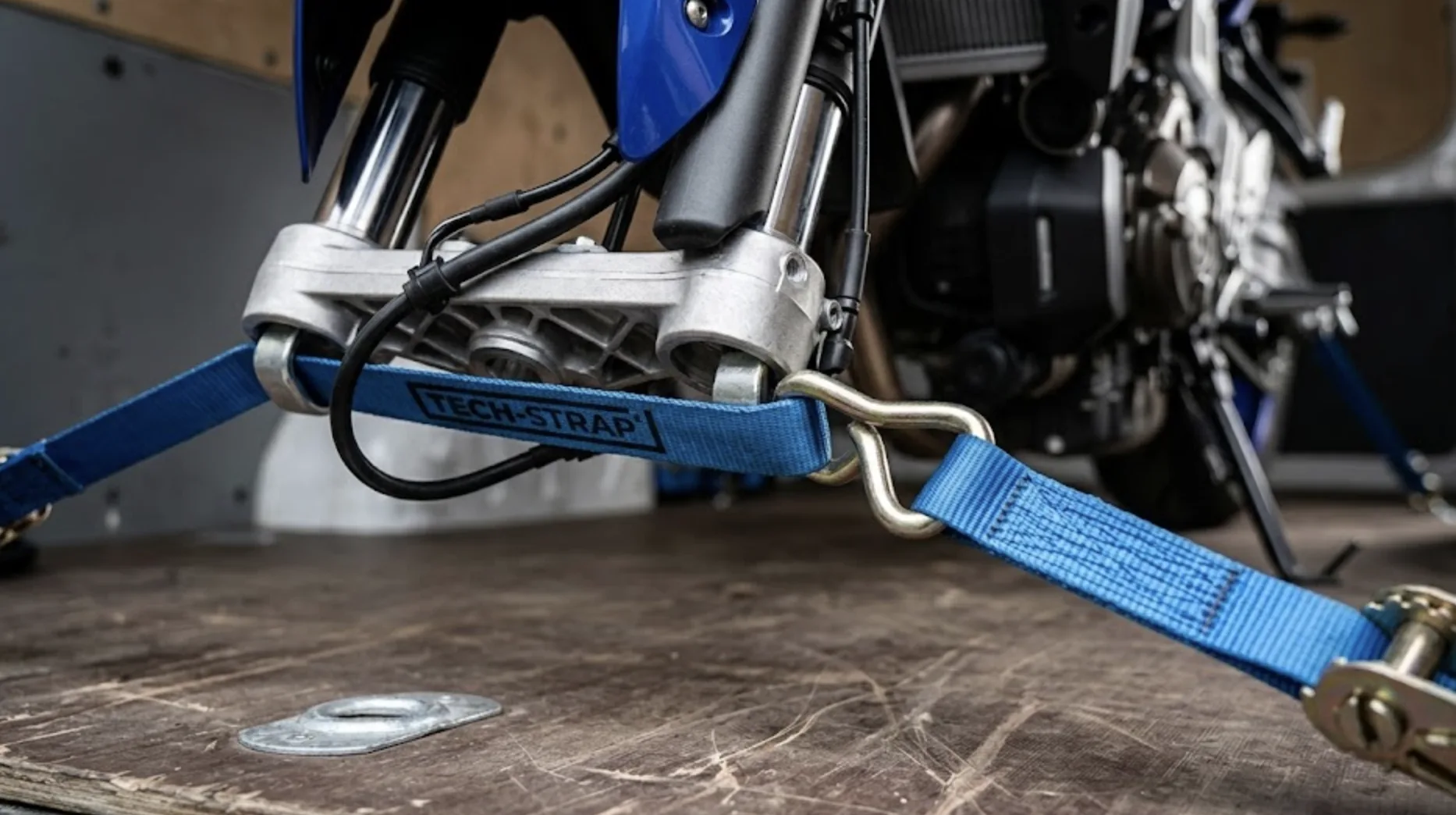 Gros plan sur une sangle à cliquet bleue fixée au té de fourche inférieur d'une moto dans un fourgon, suspension légèrement comprimée