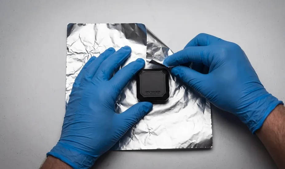 Mains avec gants de protection bleus manipulant un traceur GPS et l'enveloppant dans du papier aluminium pour couper le signal